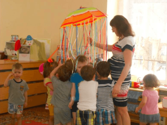 Дети и педагог держатся за разноцветные ленты, прикреплённые к куполу зонтика