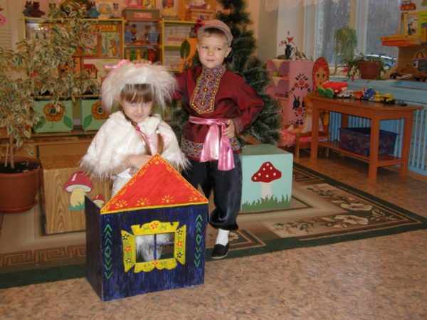 Мальчик стоит рядом с девочкой, которая прячется в картонном домике