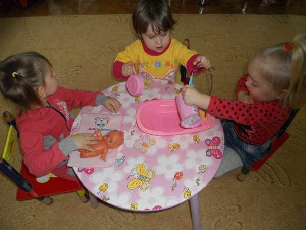 Трое детей купают куклу, сидя за круглым столиком