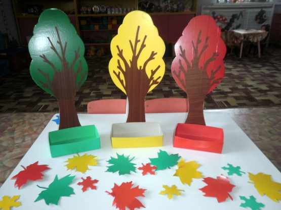 Три игрушечных дерева (красное, зелёное, жёлтое) с коробочками, много листьев этих же цветов