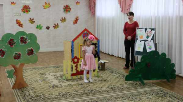 Девочка с розовым цветком на голове и воспитатель на фоне декораций — дерева, куста и домика на заднем плане