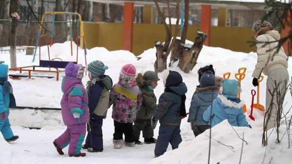 Дети и воспитательница на прогулке зимой, малыши подходят за лопатками для уборки снега
