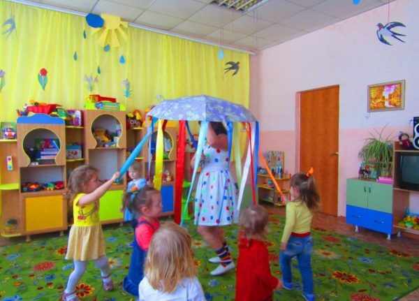 Воспитательница держит зонт, к спицам которого привязаны разноцветные ленты, за них держатся дети