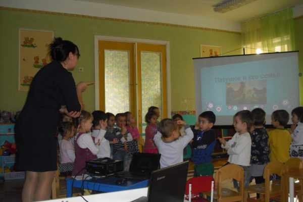 Воспитательница рукой показывает детям петушка, на заднем фоне презентация с изображением петушка и его семьи