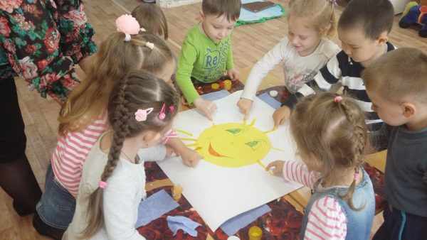 Дошкольники вместе рисуют лучики изображённому на ватмане солнышку