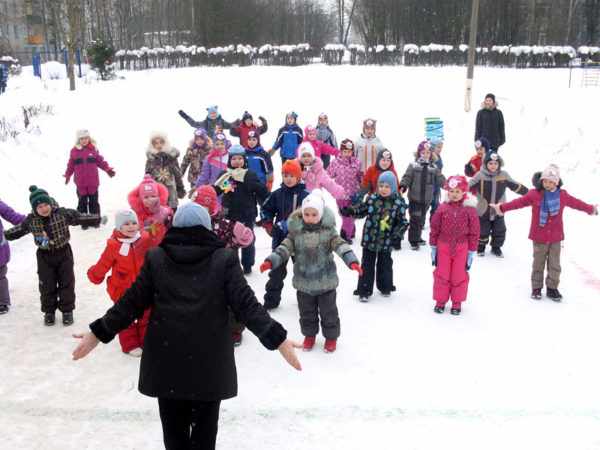 Дети в зимней одежде выполняют спортивные упражнения, воспитатель показывает