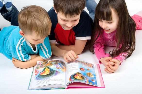 Два мальчика и девочка, лёжа на животе, читают книгу с картинками