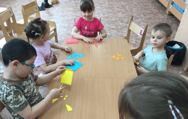 Дети, сидя за столом, собирают изображения из цветных бумажных фигур