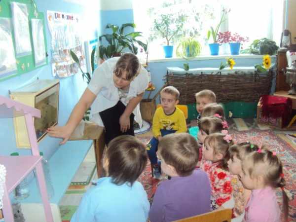 Воспитательница рассказывает о рыбах в аквариуме сидящим на стульях детям