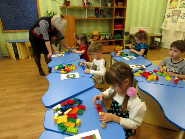 Дети за столами занимаются Лего-конструированием, воспитатель помогает