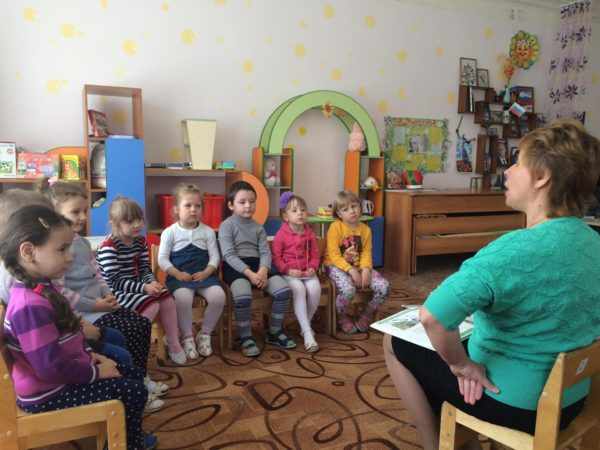 Педагог с книгой на коленях обращается к дошкольникам