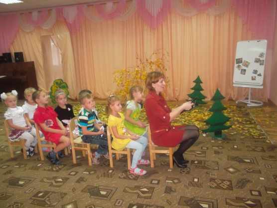 Воспитательница с детьми сидят паровозиком на стульях, на заднем плане — антураж леса