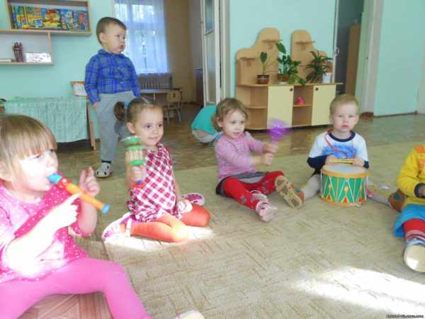 Дети, сидя на ковре, играют на различных музыкальных инструментах
