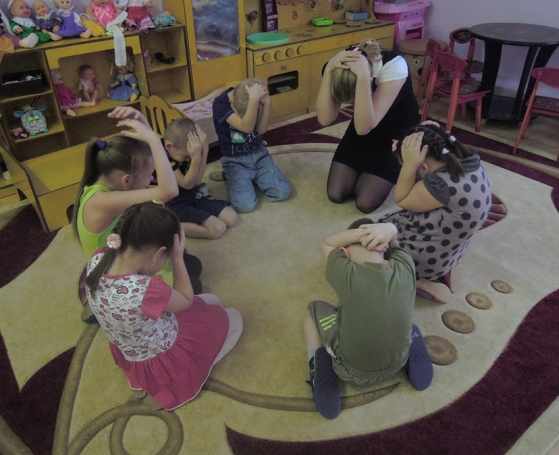 Воспитательница и дети обхватили голову руками, сидят на коленях на полу