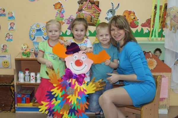 Молодая воспитательница и дети держат перед собой клоуна из бумажных детских ладошек