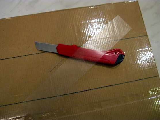 На листе картона лежат канцелярский нож и полоса прозрачного пластика