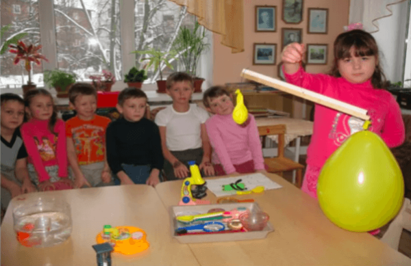 Девочка держит в руках импровизированные весы с воздушными шарами, остальные дети наблюдают