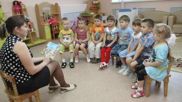 Воспитательница читает книжку детям, сидящим полукругом
