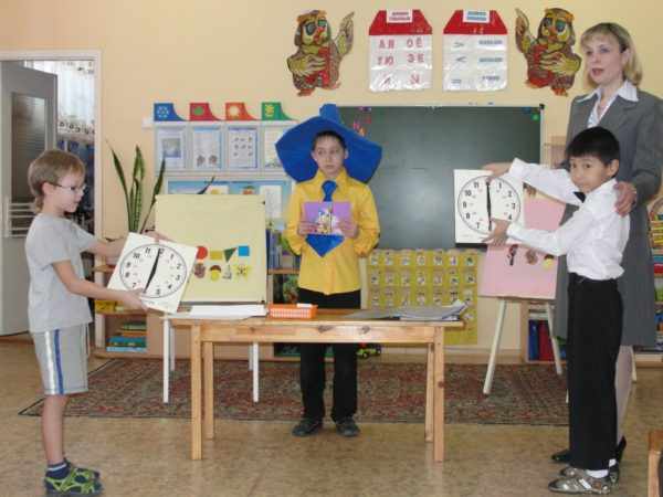 Ребёнок в костюме Незнайки, два мальчика с картонными часами в руках и педагог