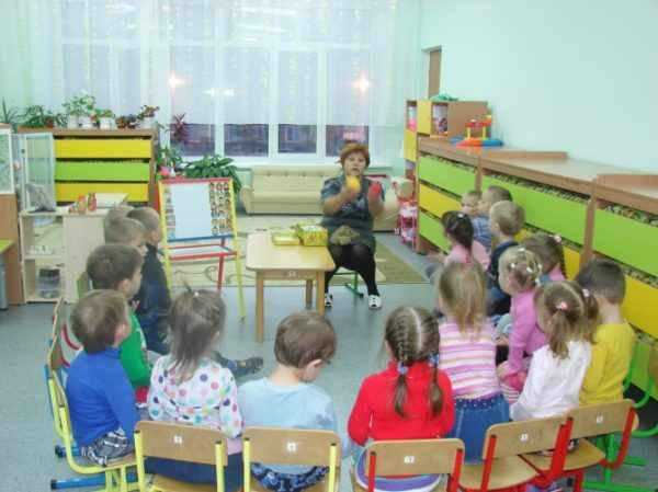 Воспитательница что-то говорит, показывая фрукты, дети слушают, сидя по кругу на стульях