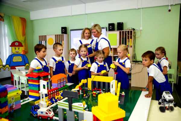 Воспитатели и дети в синих комбинезонах стоят около моделей, выполненных из конструктора