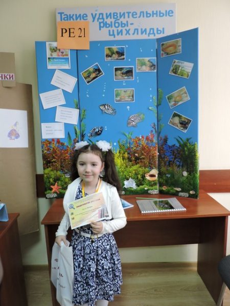 Девочка с грамотой в руках стоит на фоне стенда с фотографиями редких рыб