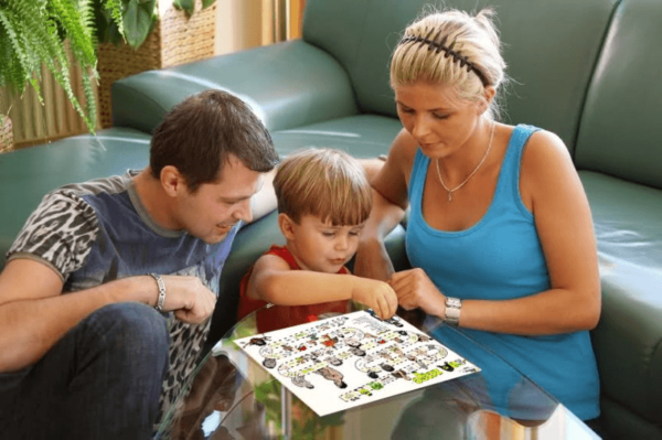 Мальчик вместе с мамой и папой размещает наклейки на листе, лежащем на стеклянном столике