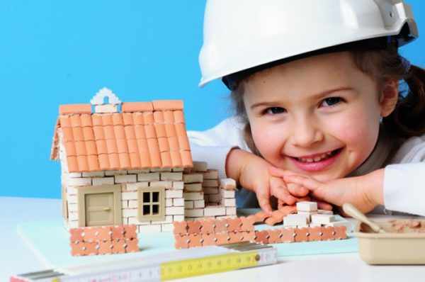Девочка в строительной каске рядом с недостроенным игрушечным домиком