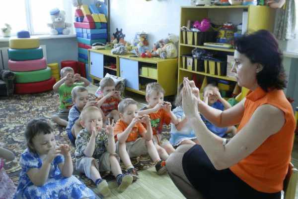 Воспитательница с детьми, сидящими на коврике, проводит пальчиковую гимнастику