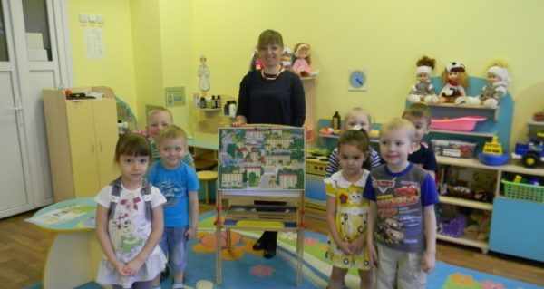 Воспитательница придерживает мольберт с картинкой города, дети стоят по обе стороны от неё