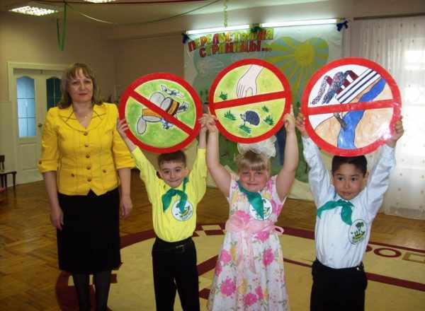 Трое детей держат запрещающие знаки по правилам поведения на природе, воспитатель стоит рядом