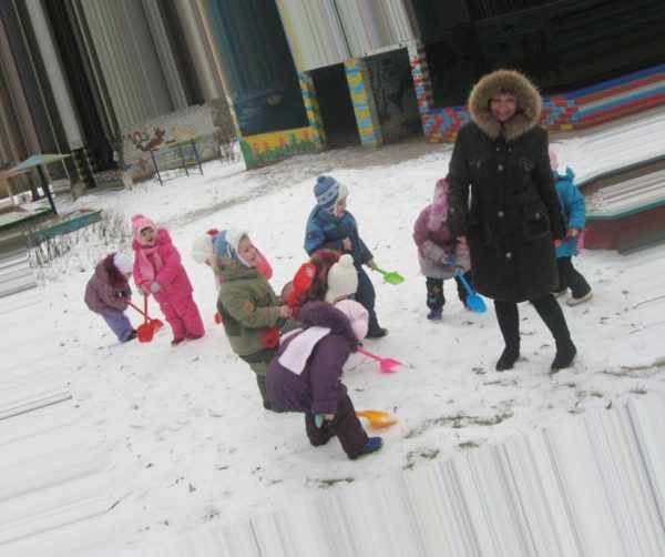 Дети во дворе с помощью лопаток убирают снег, воспитатель улыбается