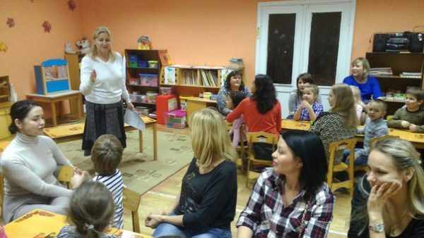Воспитательница выступает перед сидящими по группам родителям с детьми