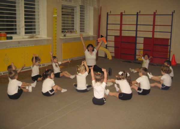 Воспитательница проводит физкультурное занятие в спортзале, дети сидят, вытянув руки вверх