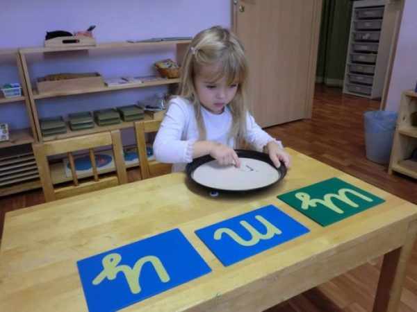 Девочка рисует буквы на песке, рядом на столе — шершавые буквы