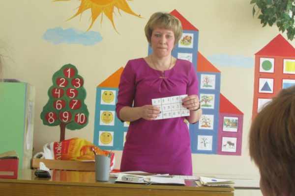 Воспитательница стоит с карточкой в руках на фоне бумажного дерева с цифрами и домиков из цветной бумаги на стене