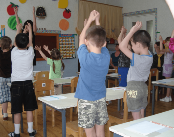 Дети с поднятыми вверх руками делают гимнастику возле парт