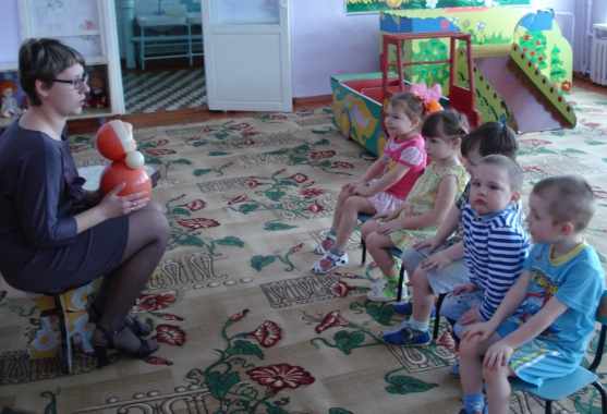 Воспитательница показывает матрёшку детям, сидящим на стульях