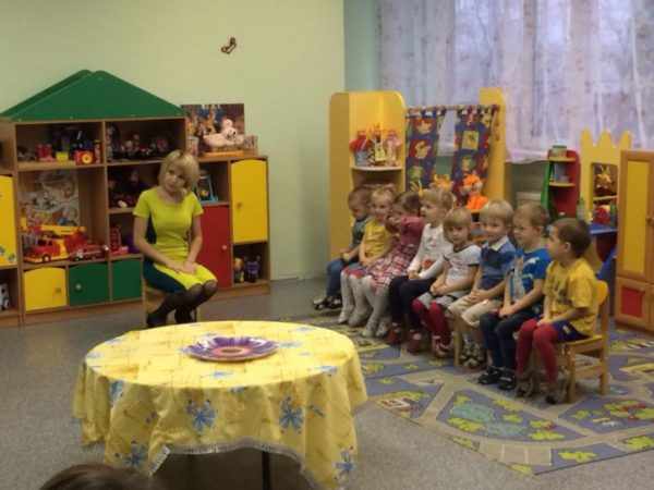 Дети сидят на стульях, воспитательница смотрит в сторону зрителей, на переднем плане стол, застеленный скатертью