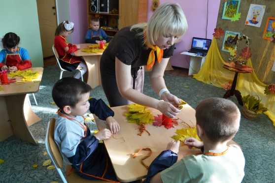 Педагог показывает детям, сидящим за столом, осенние листья