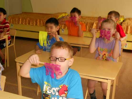 Дети сидят за столами и дуют на розовые и жёлтые флажки, на переднем плане мальчик в голубой футболке с Angry Birds