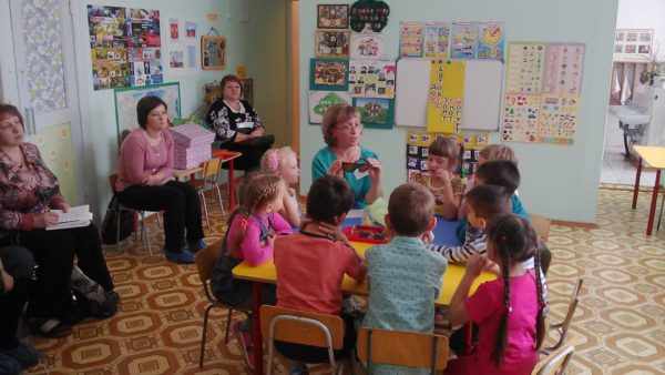 Воспитательница сидит вместе с детьми за круглым столом, взрослые сидят по периметру комнаты