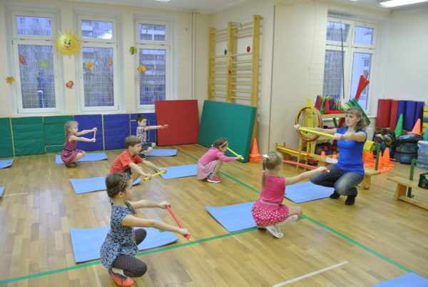Дети и педагог выполняют приседания с гимнастическими палками в руках