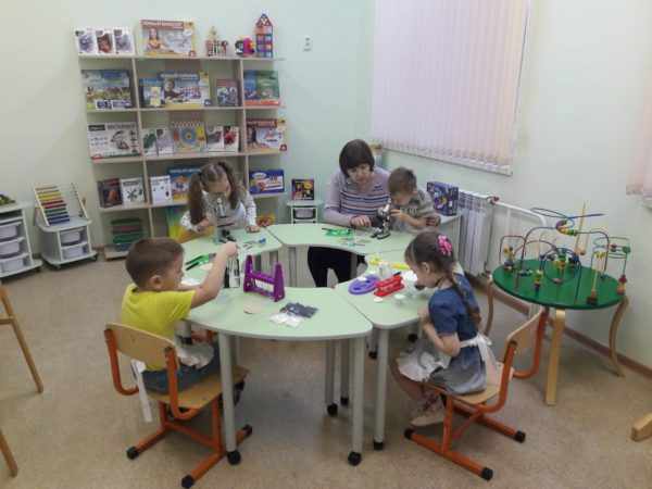 Дети, сидя за столами, работают с микроскопами, педагог помогает