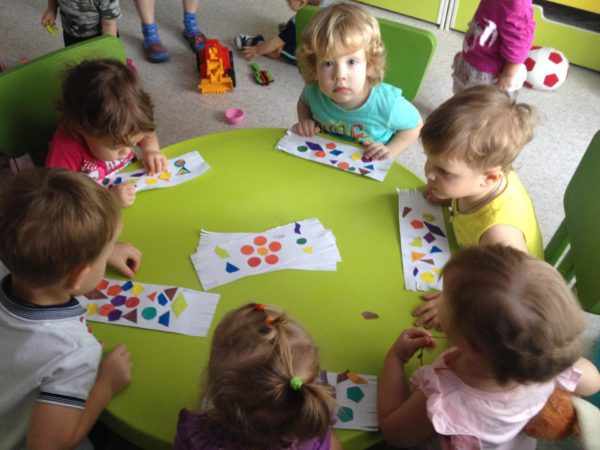 Дети, сидя за столом, составляют узоры на бумаге из разноцветных геометрических фигур