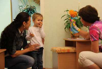 Ребёнок стоит рядом с мамой в дверях, воспитательница показывает ему куклу