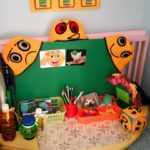 Центр психологической разгрузки:жёлтый стол полукругом с материалами на нём и оранжевыми вязаными шапочками с эмоциями на зелёной доске